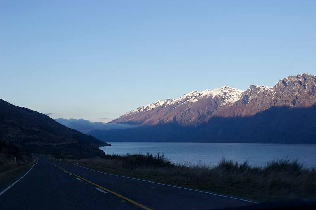 Driving to Manapouri with the road to the left and the river to the right with beautiful snow-capped mountains in the background in New Zealand