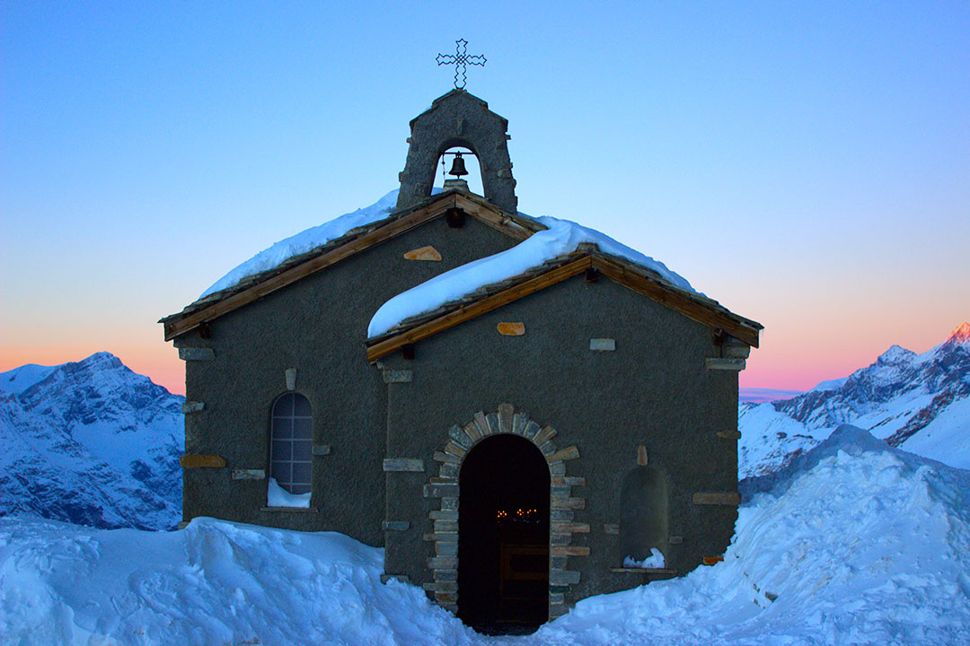 Beautiful little church covered in snow with candles lit up inside and the orange and pink sunset in the background surrounded by snow