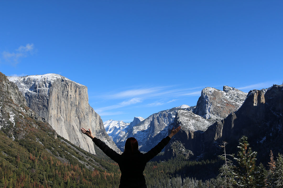 Me standing in the foreground facing away with my hands in the air looking over snowy El Capitan and Yosemite National Park on a beautiful blue sky winter day