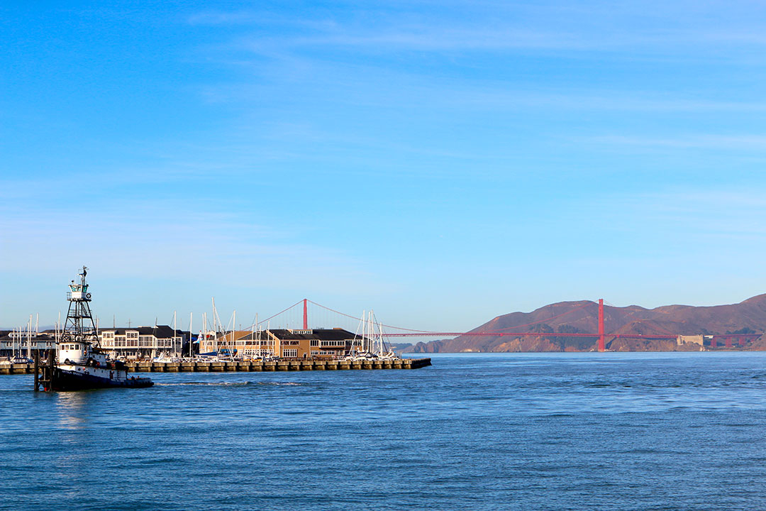 View of the Golden Gate Bridge in the distance beyond a pier with mountains in the background on a bright blue sky day