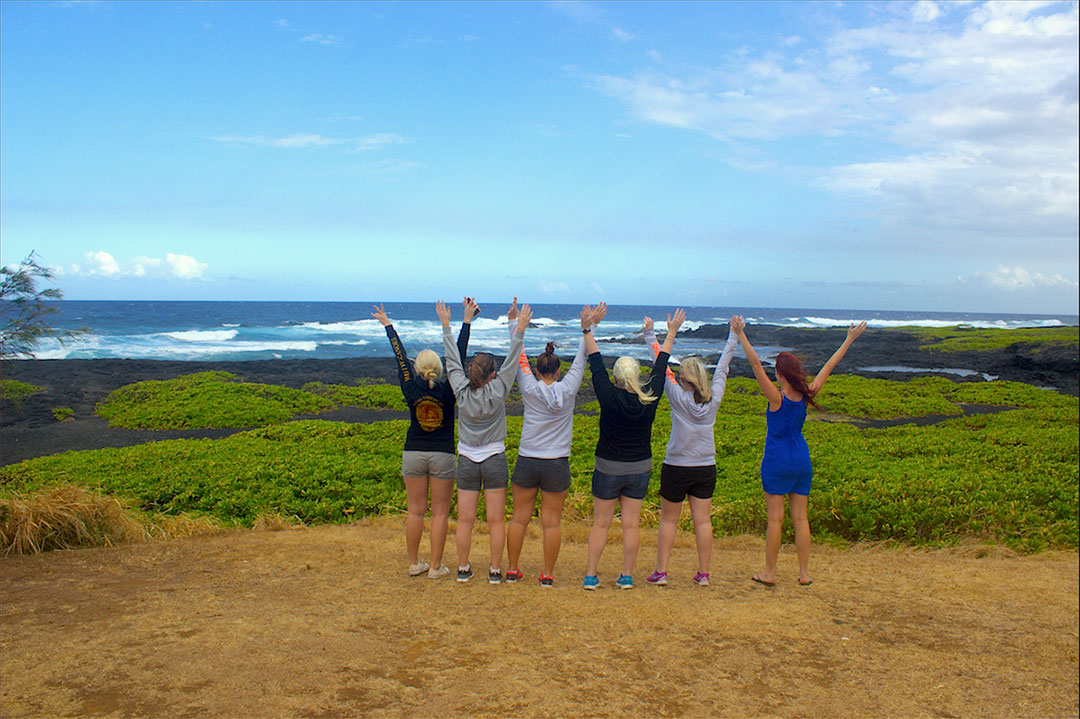 A group of 6 girl friends facing a rough black sand beach in Hawaii with hands in the air as the wind blows our hair