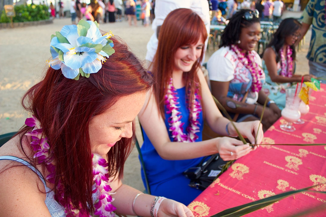 Hollie and I weaving a head-dress out of long thin green leaves while smiling and wearing leis around our neck and flowers in our hair