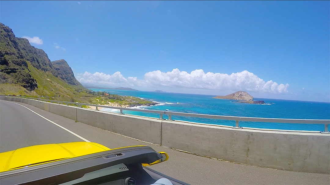 View from the yellow Chevrolet Camaro with the top down looking out over the bright blue ocean and rugged mountains while on a drive around Oahu