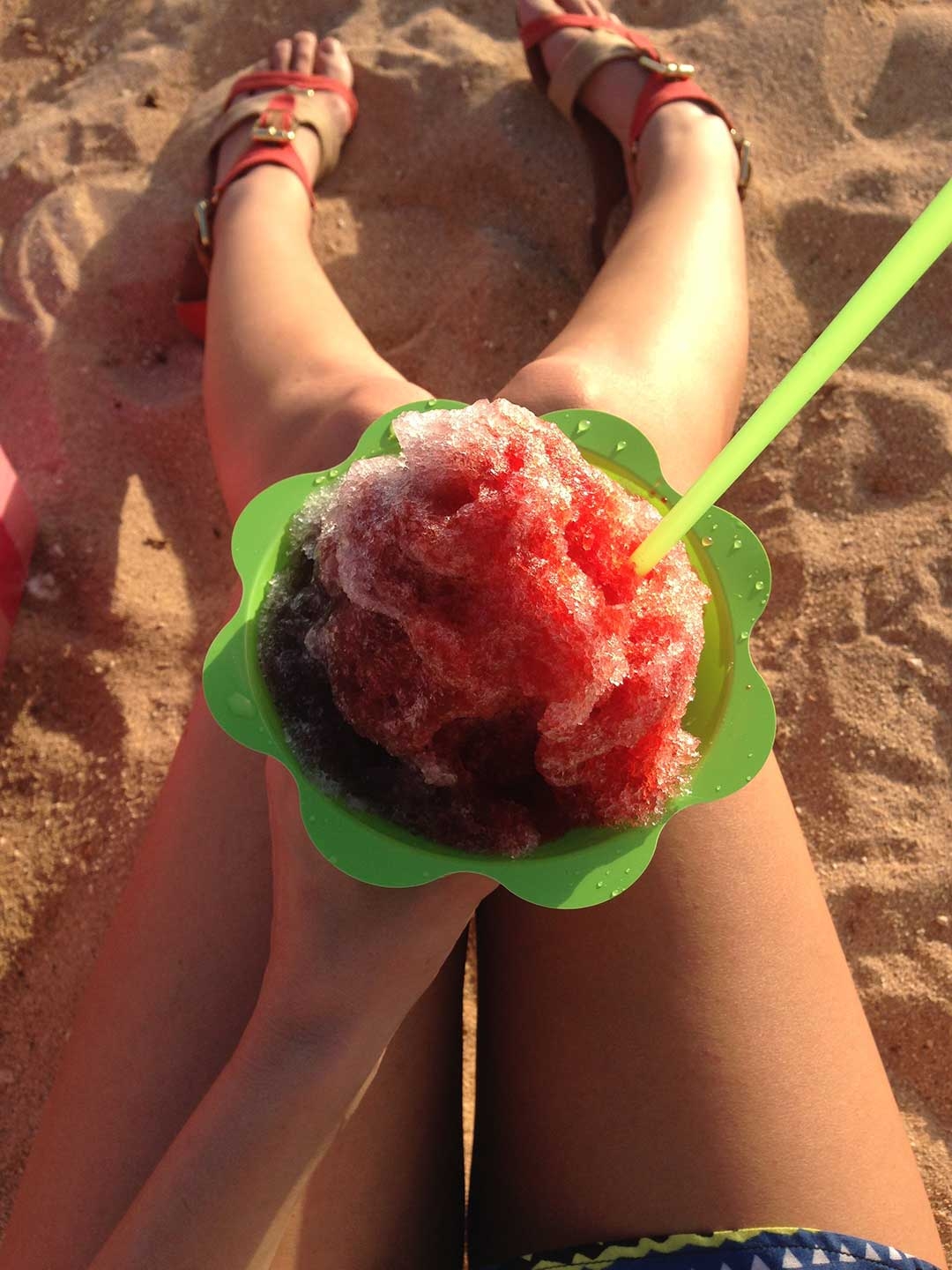 Red and blue shaved ice in a green cup resting on my legs, being enjoyed on Waikiki beach, Hawaii