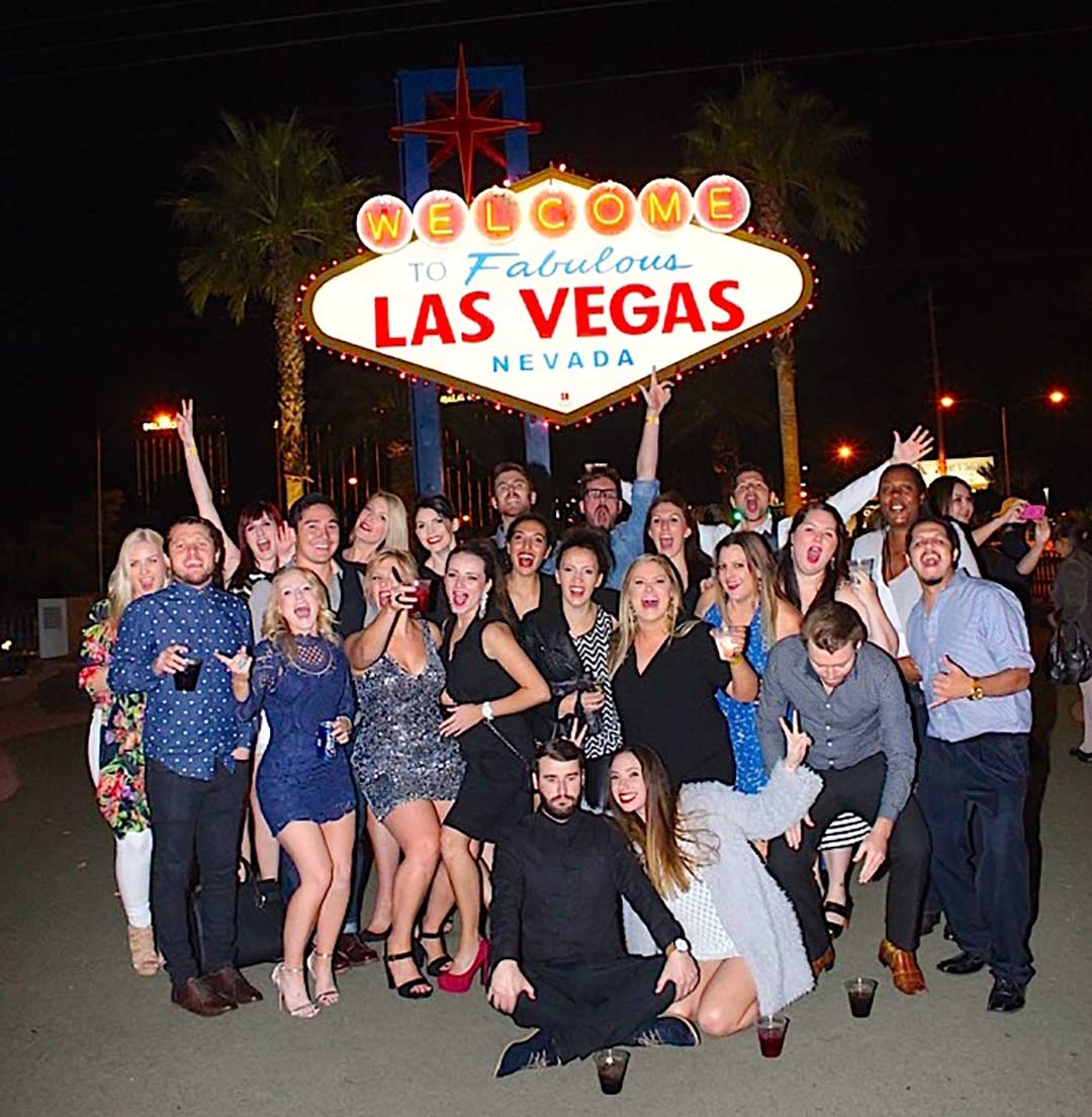 Group of friends in smiling and waving in front of the Welcome to Fabulous Las Vegas sign at night
