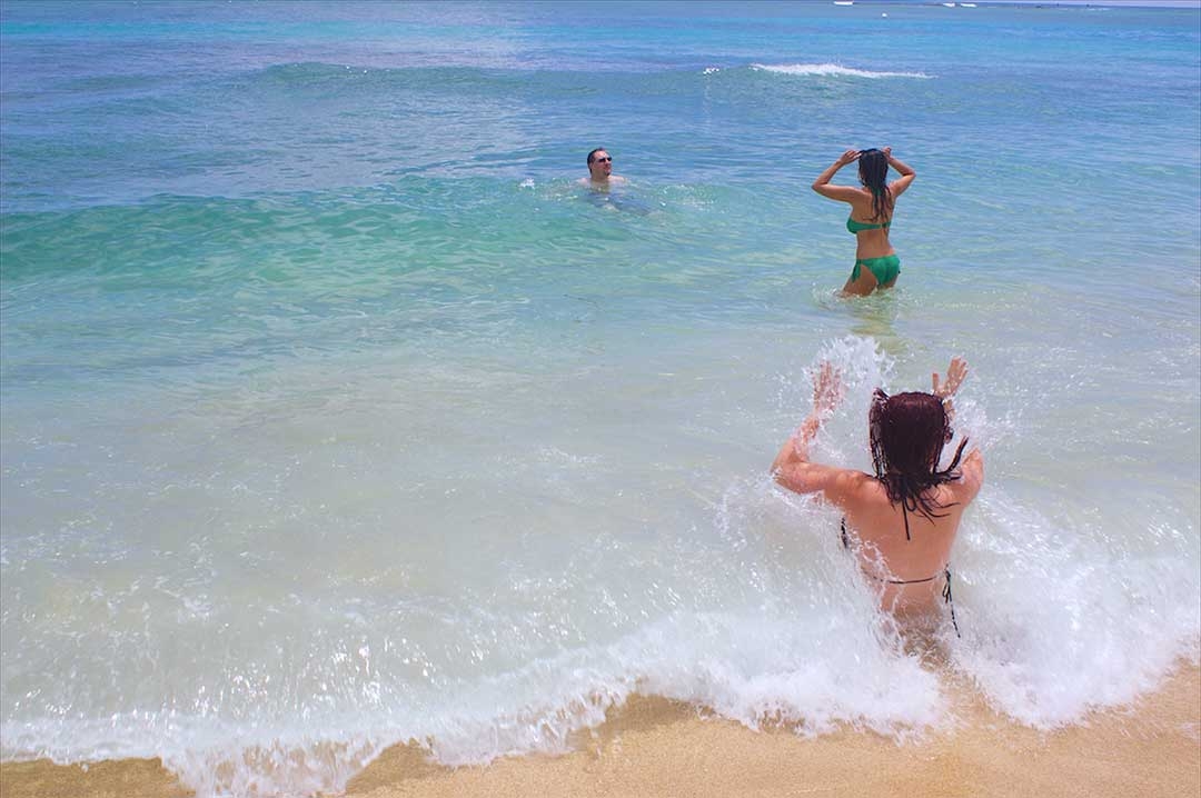 Girl sitting in the shallows on a beautiful beach while a small wave crashes in her face and she unsuccessfully puts her hands up to protect her self from the splash