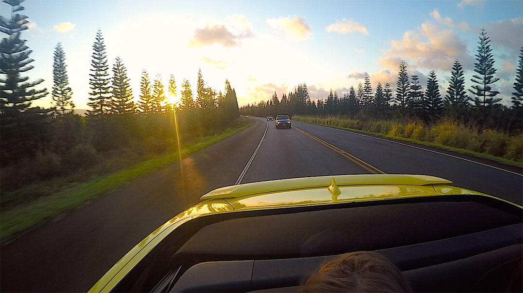 View of the Hawaiian sunset out the back of the Chevrolet Camaro with the top down while on a drive down a road surrounded by tall trees