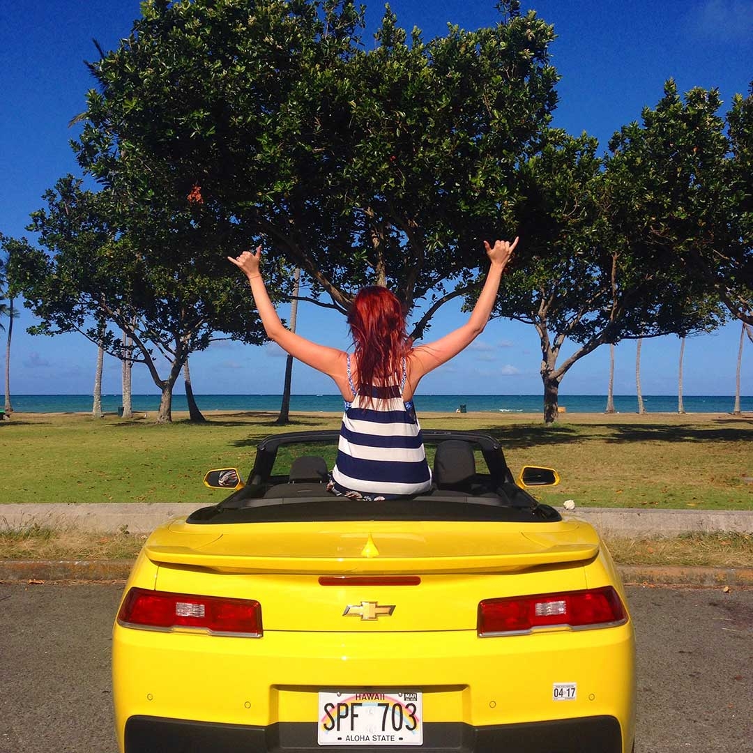 Yellow Chevrolet Camaro parked in the carpark in front of a beautiful Hawaiian beach and green trees with me sitting on the back facing the ocean with my hands in the air