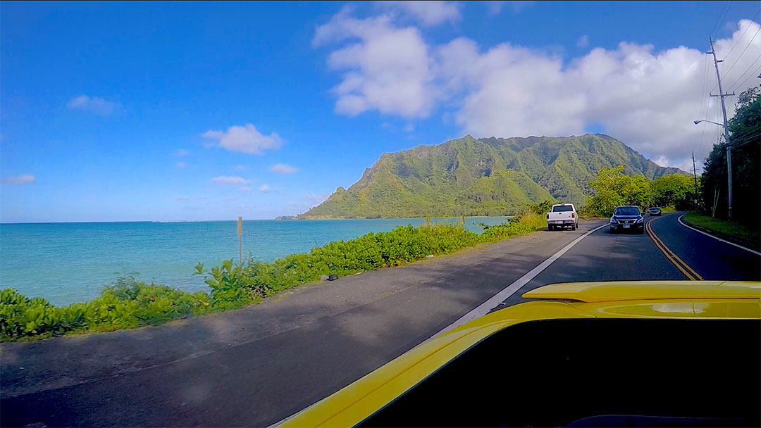 View from the yellow Chevrolet Camaro with the top down looking out over the bright blue ocean and rugged mountains while on a drive around Oahu