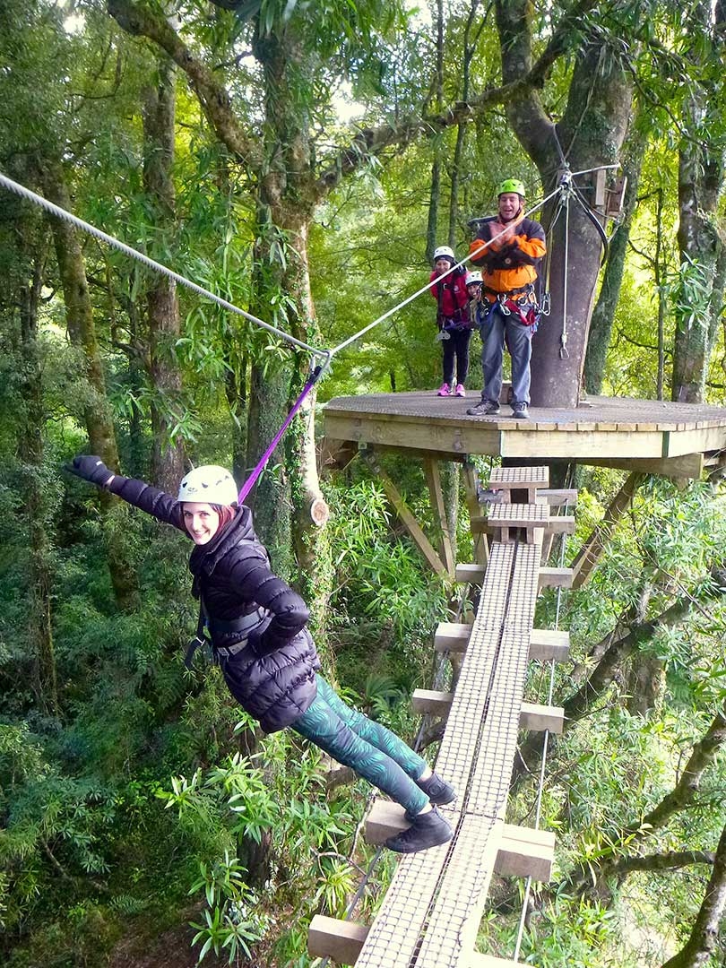 Me doing a superman pose while hanging off the edge of a swing bridge without railings with the tour guide clapping on the treetop platform in the background