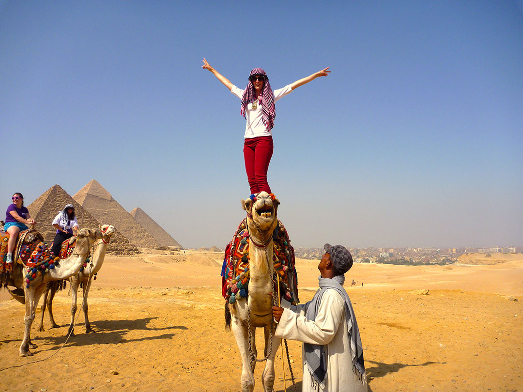 Me standing on the back of Charlie the camel with my hands in the air after racing around the desert in front of the Grand Pyramids of Giza and some other members of the tour group